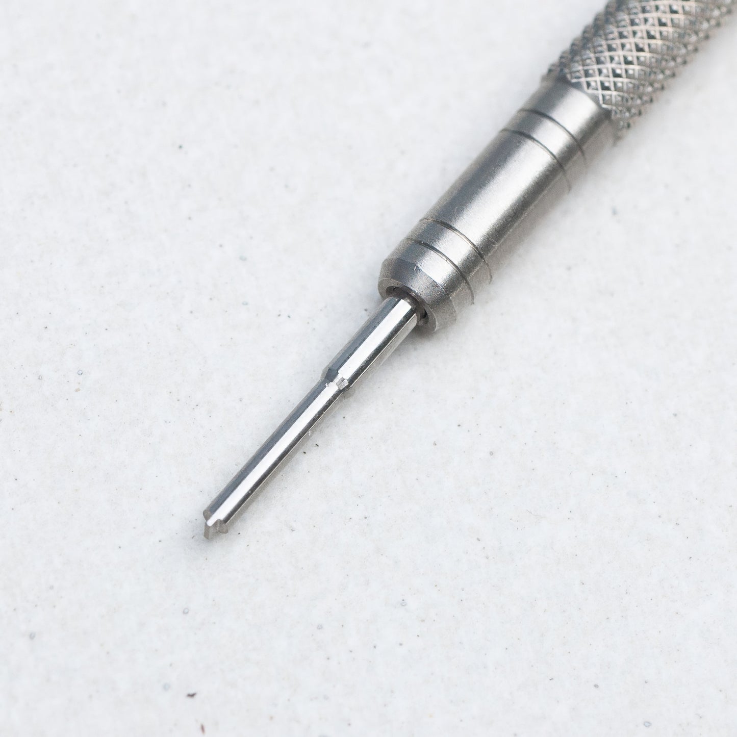A close-up of a silver metal precision screwdriver bit with a knurled textured handle, shown on a light gray textured background.