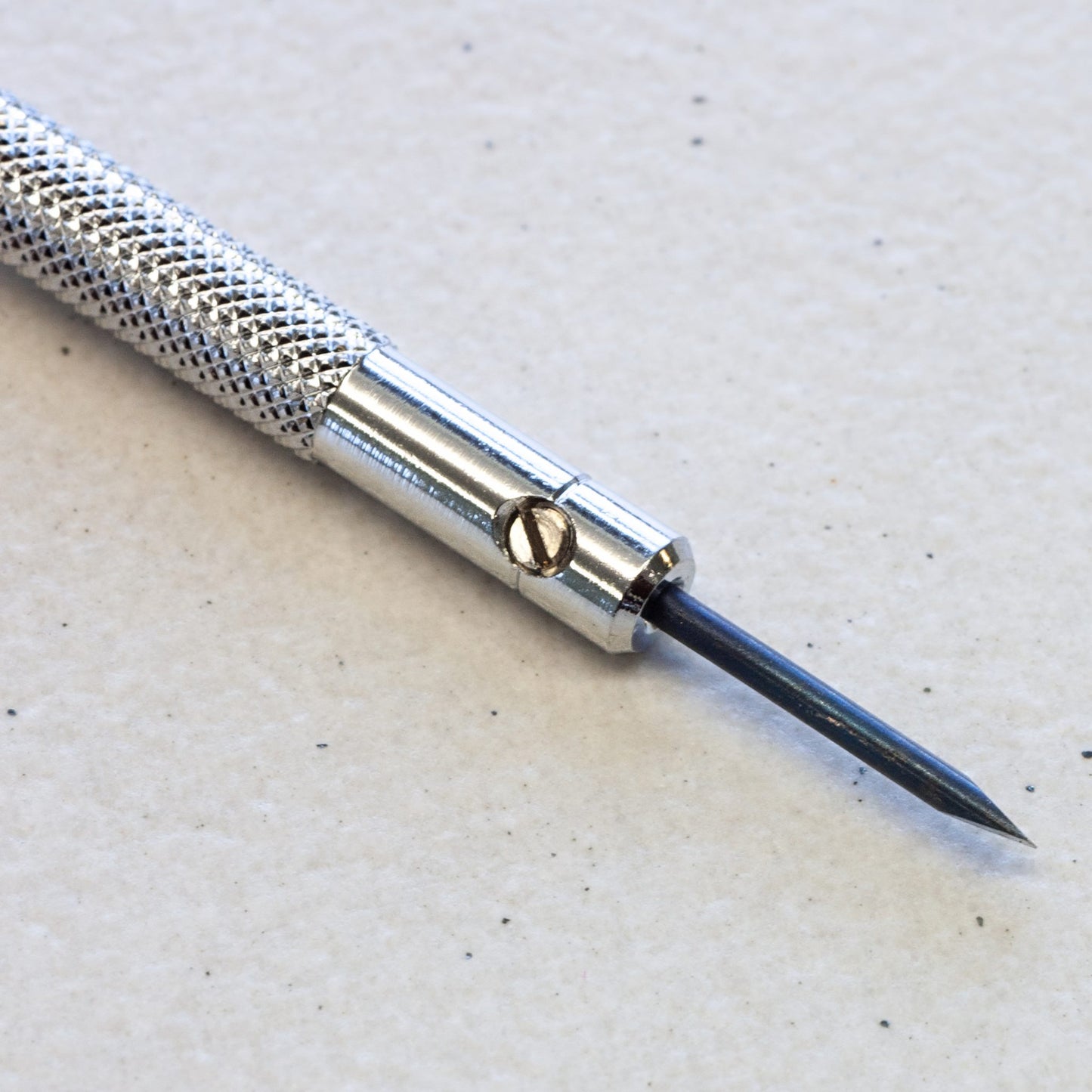 A close-up of a precision craft tool with a textured silver handle, a polished metal collar, and a sharp dark blue or black pointed tip, resting on a light textured surface.