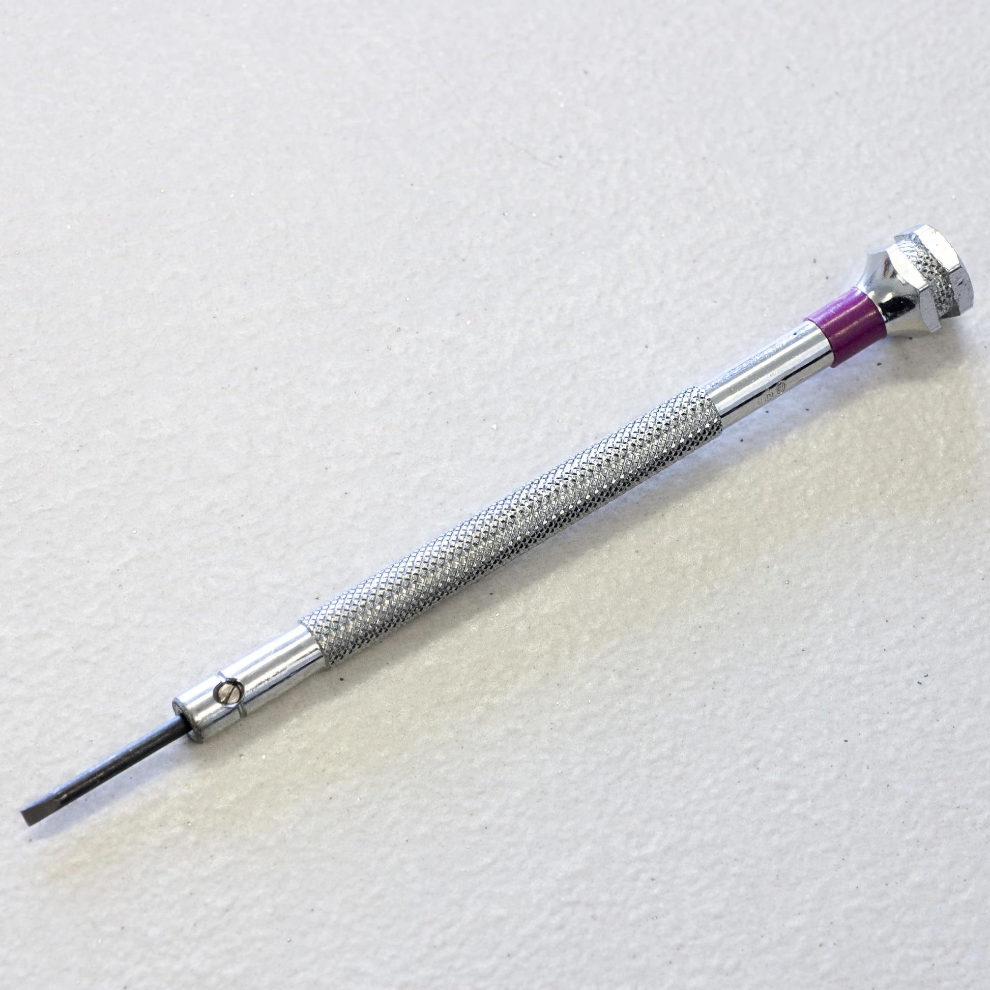 A precision screwdriver with a textured silver metal handle and a purple accent ring, shown diagonally on a light textured background.