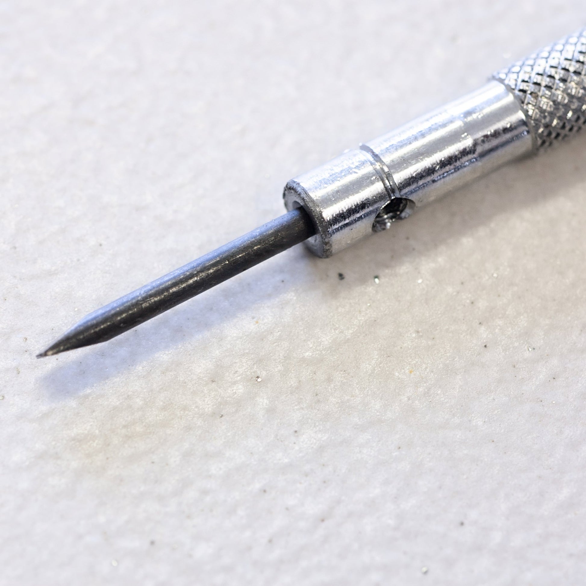 A close-up of a precision screwdriver bit with a textured silver handle and a dark metal tip, resting on a light textured surface.