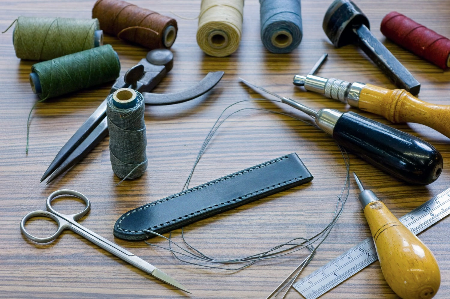 A flat-lay arrangement of leatherworking tools and spools of thread on a wooden surface, including a black leather loop, scissors, needles, wooden-handled tools, and multiple colored thread spools.