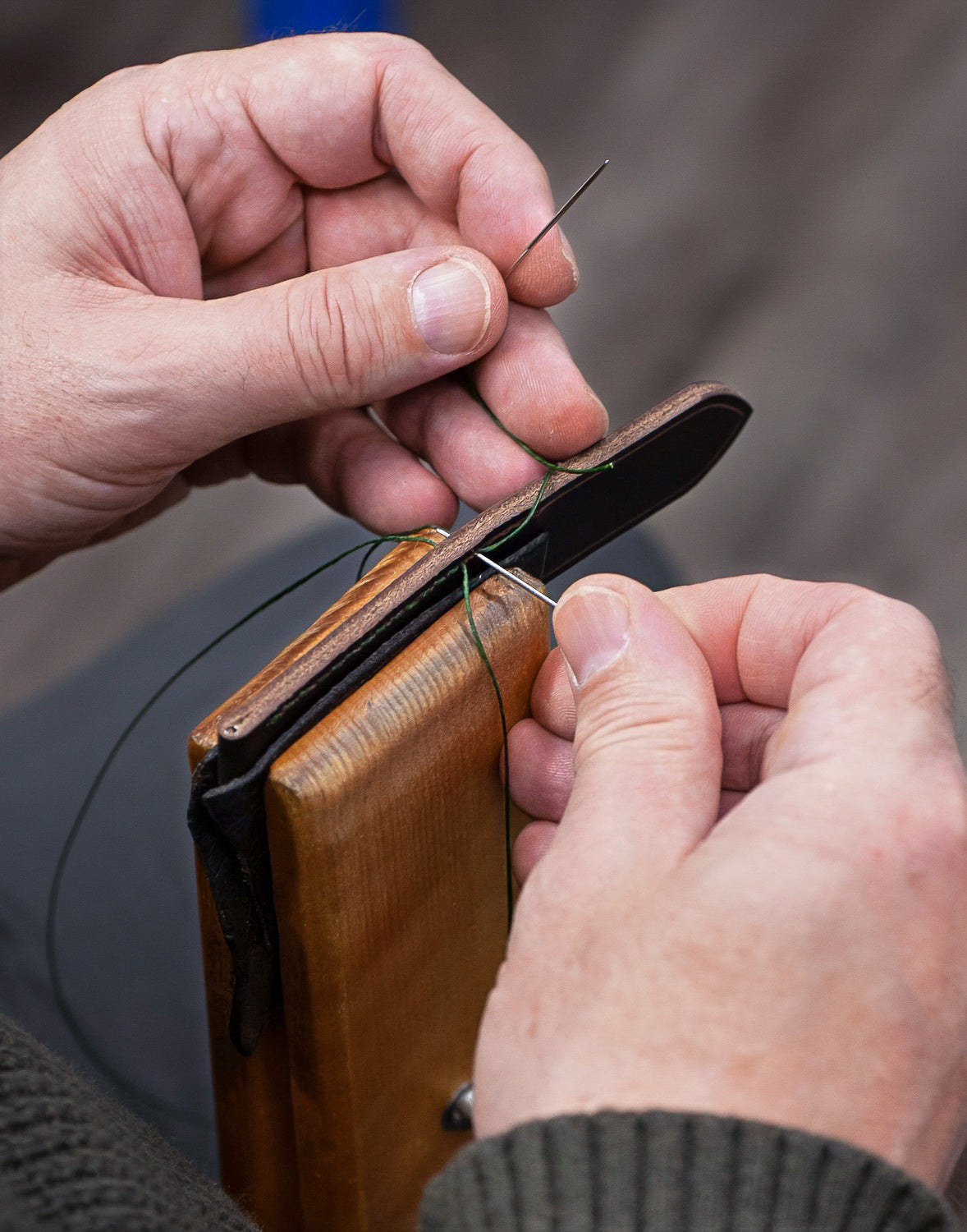 Close-up of a person's hands sewing or stitching a brown leather or wood object with a needle and green thread.