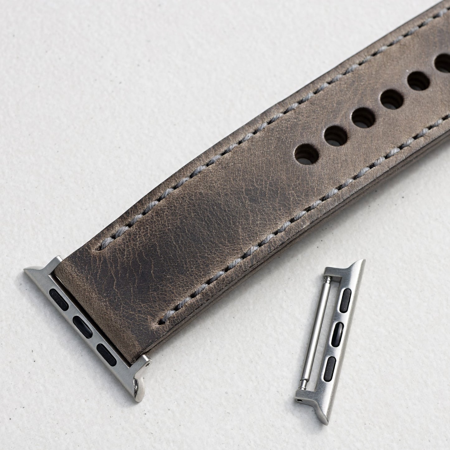 A close-up of a grey leather watch strap with silver metal hardware, on a light textured background.