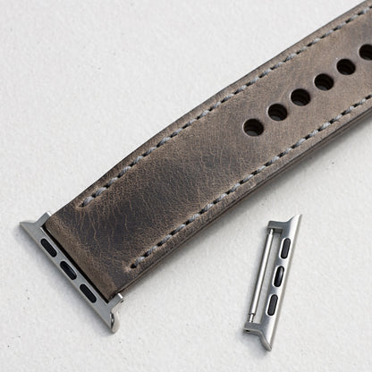 A close-up of a grey leather watch strap with silver metal hardware, on a light textured background.