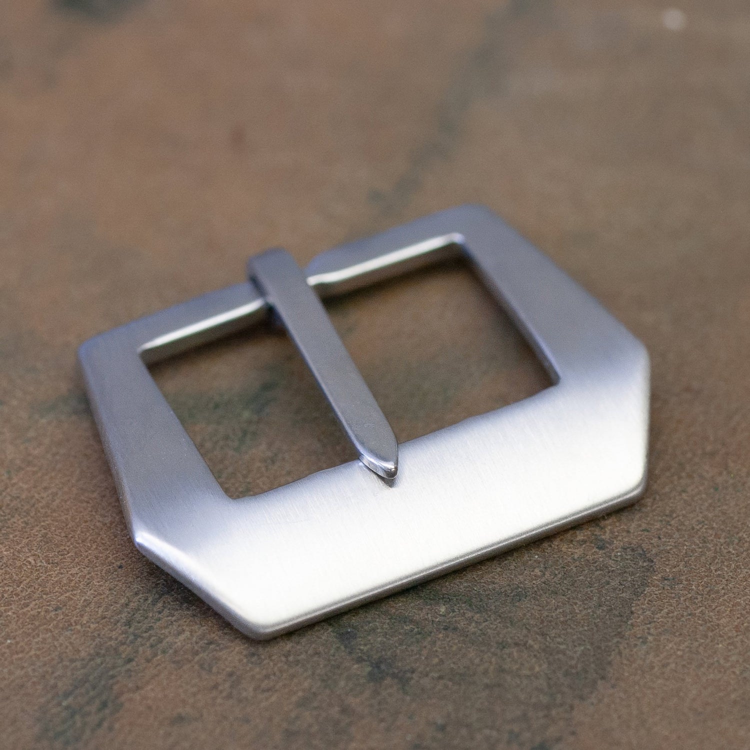 A brushed silver metal sewn in watch strap buckle with a square-shaped frame and a single prong, resting on a textured brown surface.