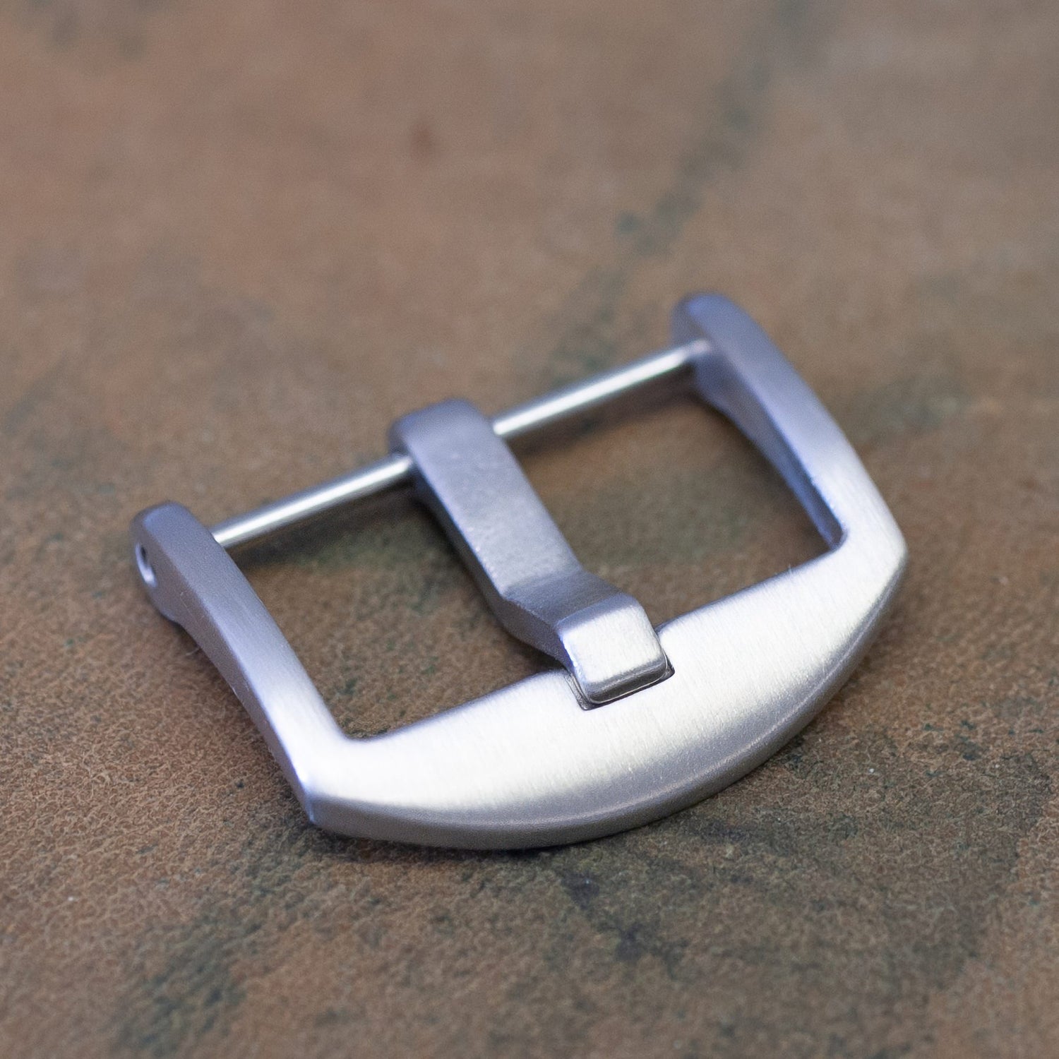 A brushed silver metal watch buckle resting on a textured brown surface.