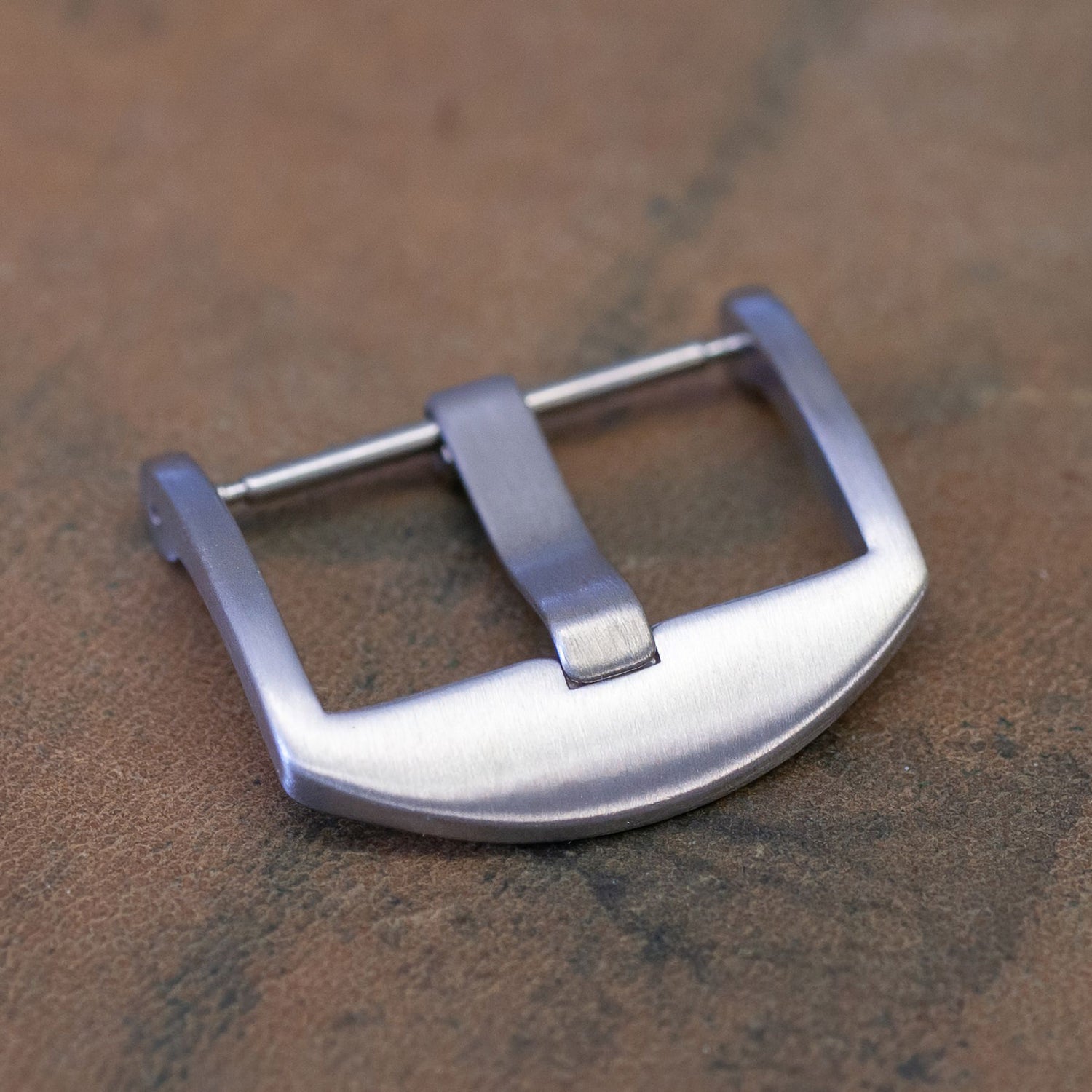 A brushed silver metal watch strap buckle resting on a textured brown surface.