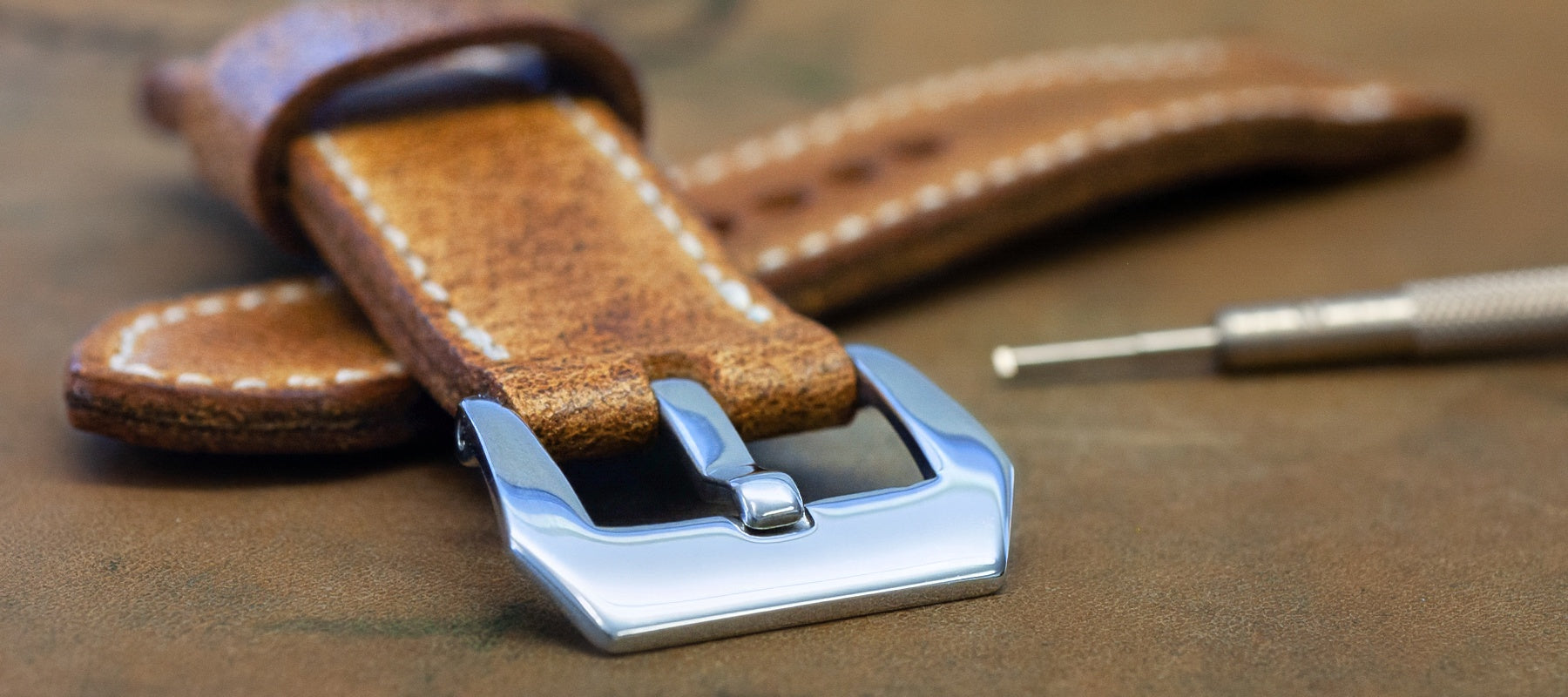 A close-up of a brown leather watch strap with white stitching and a polished silver-tone buckle, resting on a brown textured surface with a small metal tool nearby.