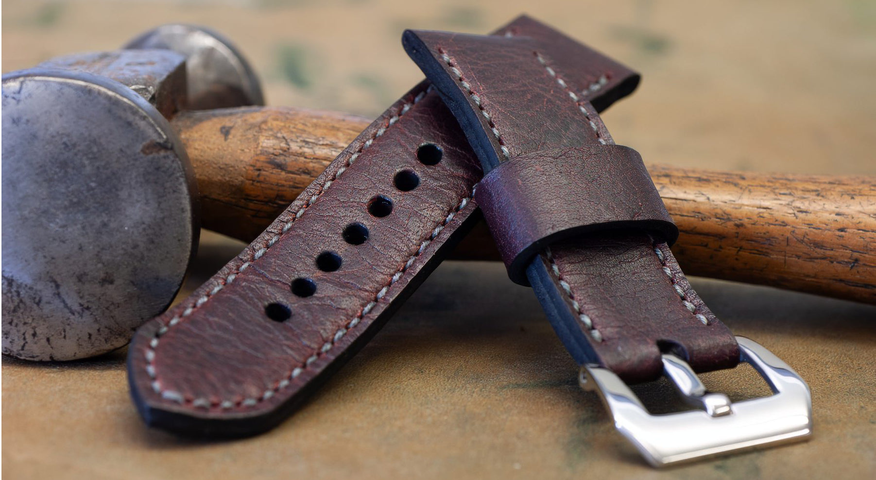 A close-up of a burgundy leather watch strap with a silver buckle, resting on a wooden tool with a metal hammer head in the background.
