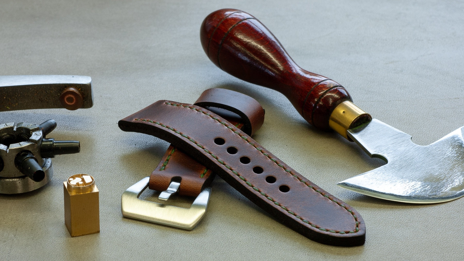 A leather watch strap with a dark brown finish and silver-toned buckle, displayed alongside a polished reddish-brown wooden handled knife and a silver metal tool on a neutral gray background.