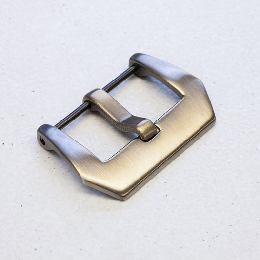 A brushed metal watch strap buckle with a warm bronze finish, photographed on a light textured background.