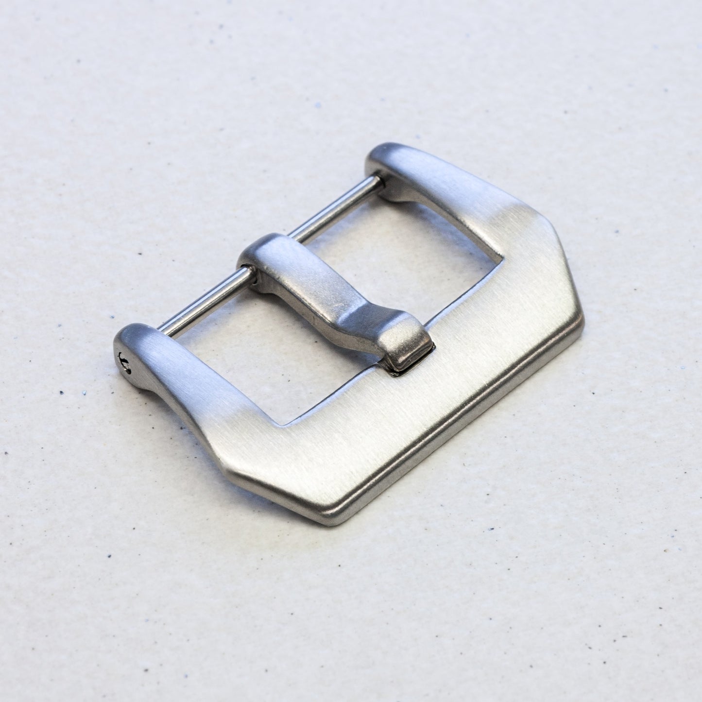 A brushed silver metal watch strap buckle, shown at an angle on a light gray textured background.