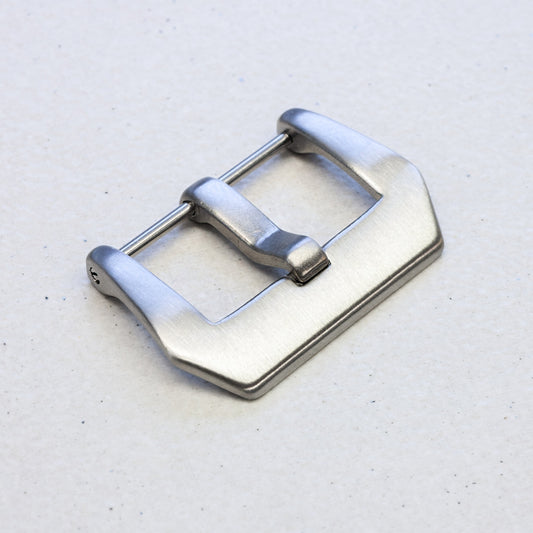 A brushed silver metal watch strap buckle, shown at an angle on a light gray textured background.