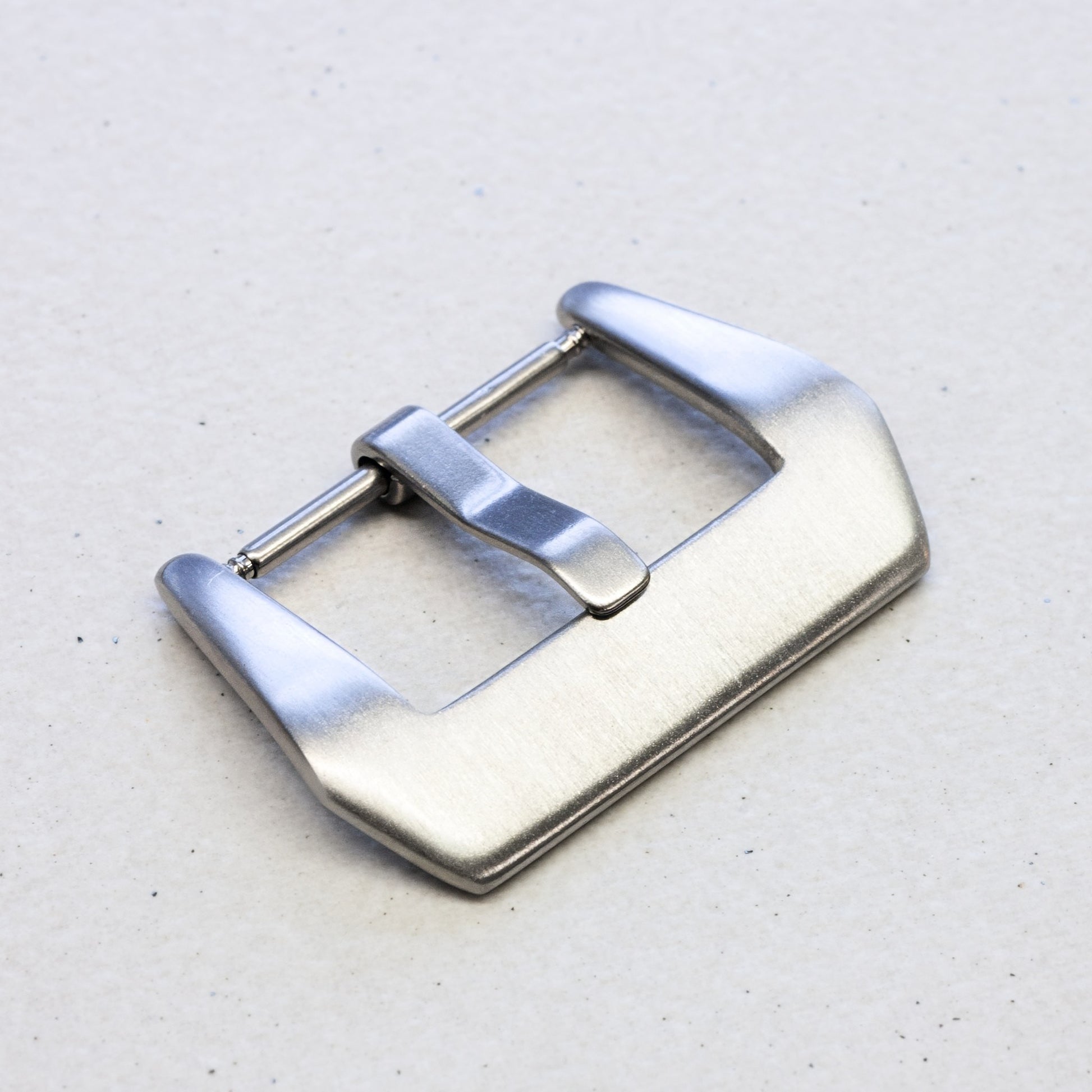 A brushed stainless steel watch strap buckle with a polished pin, shown on a light textured gray background.