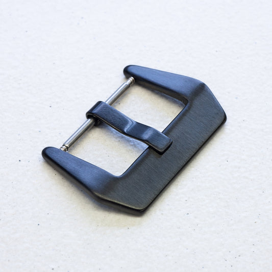 A black metal watch buckle with a brushed finish and a silver-toned pin, shown on a light textured background.