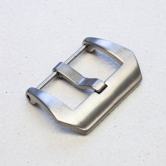 A brushed titanium watch strap buckle, isolated on a light off-white textured background.