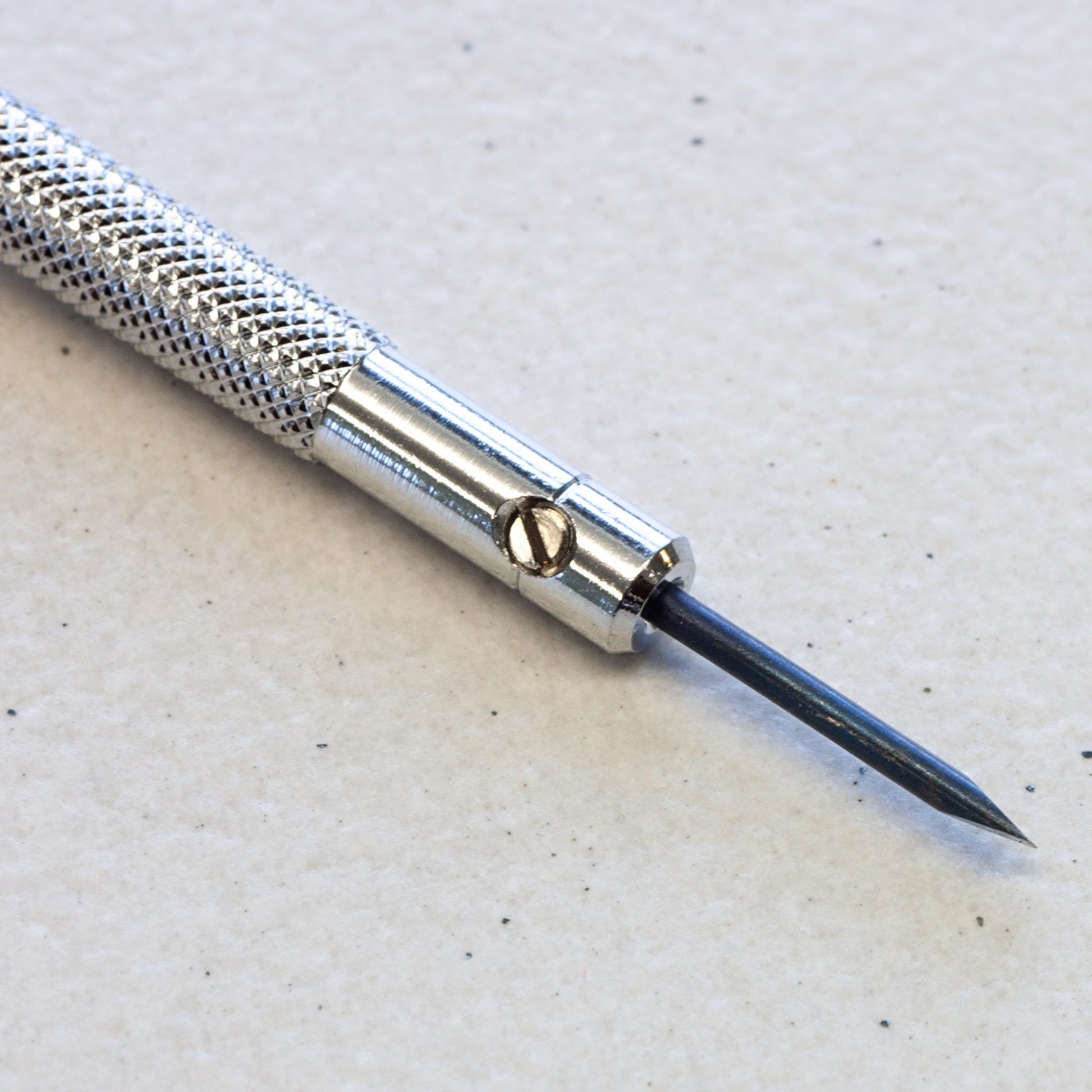 A close-up of a precision craft tool with a textured silver handle, a polished metal collar, and a sharp dark blue or black pointed tip, resting on a light textured surface.