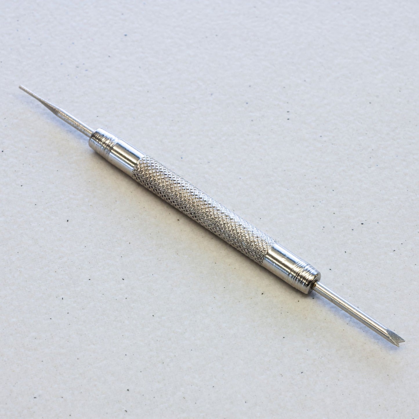 A silver metal double-ended spring bar tool with a textured knurled handle, shown on a light gray textured background.