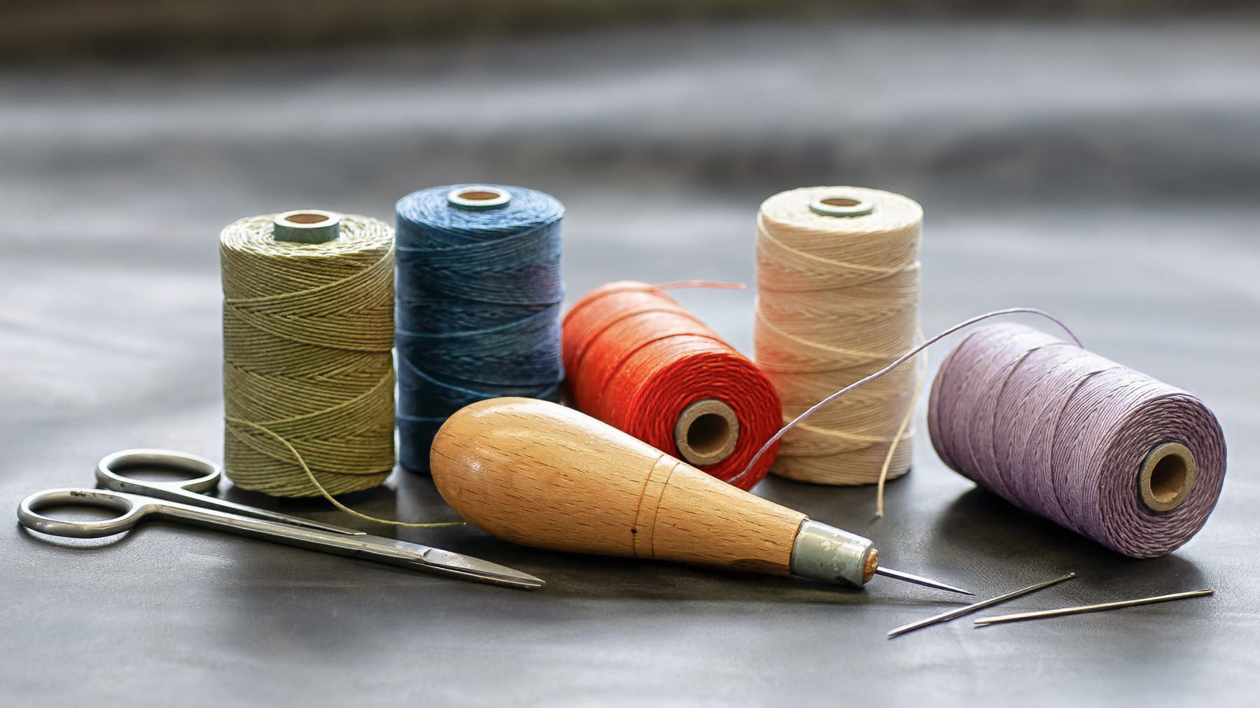 Spools of colourful linen thread and a stitching awl on a gray surface