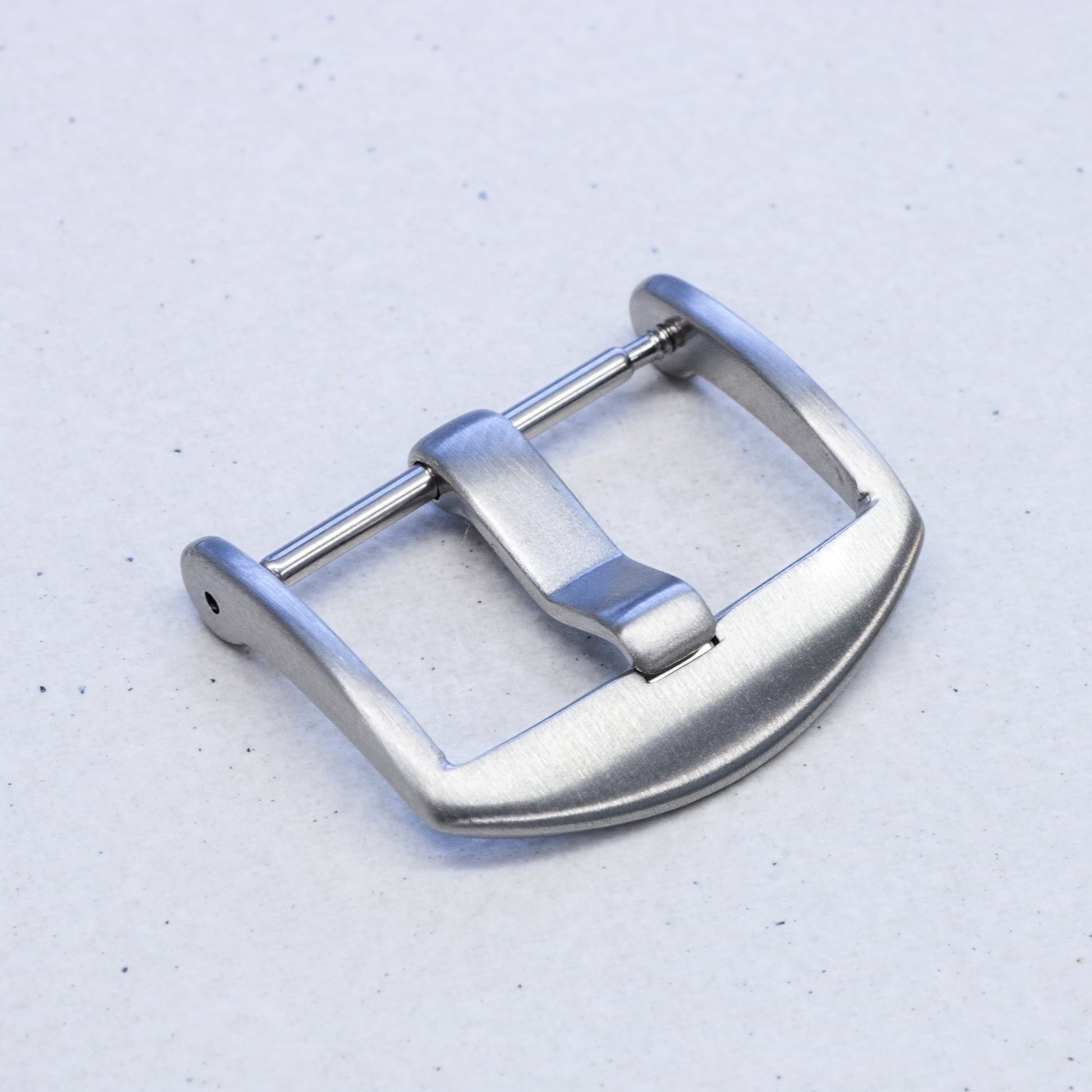 A brushed silver metal watch strap buckle, shown from a top-down angle on a light gray textured background.