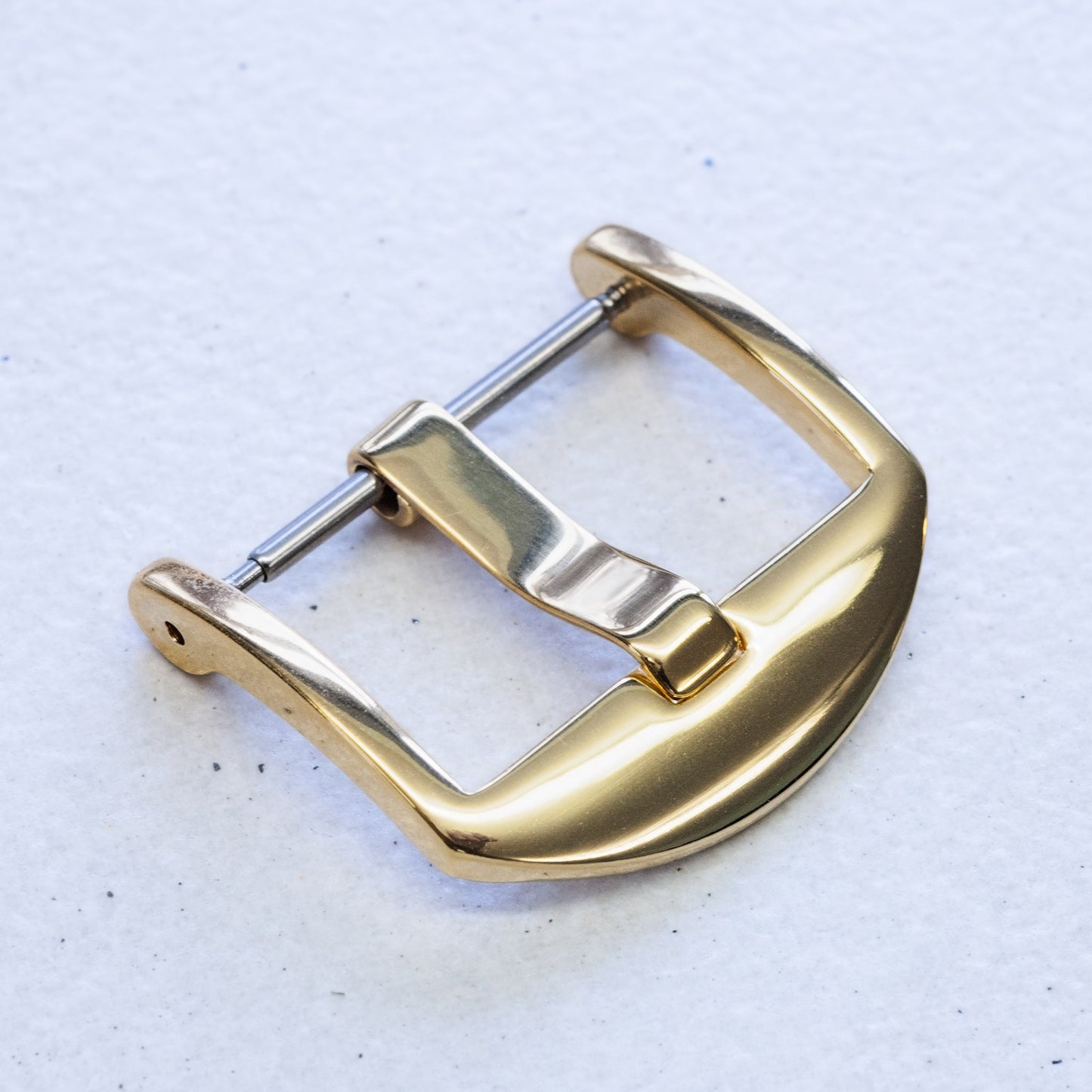 A polished gold watch strap buckle with a silver-toned pin, shown on a textured off-white background.