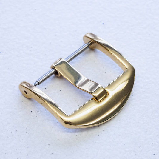 A polished gold watch strap buckle with a silver-toned pin, shown on a textured off-white background.