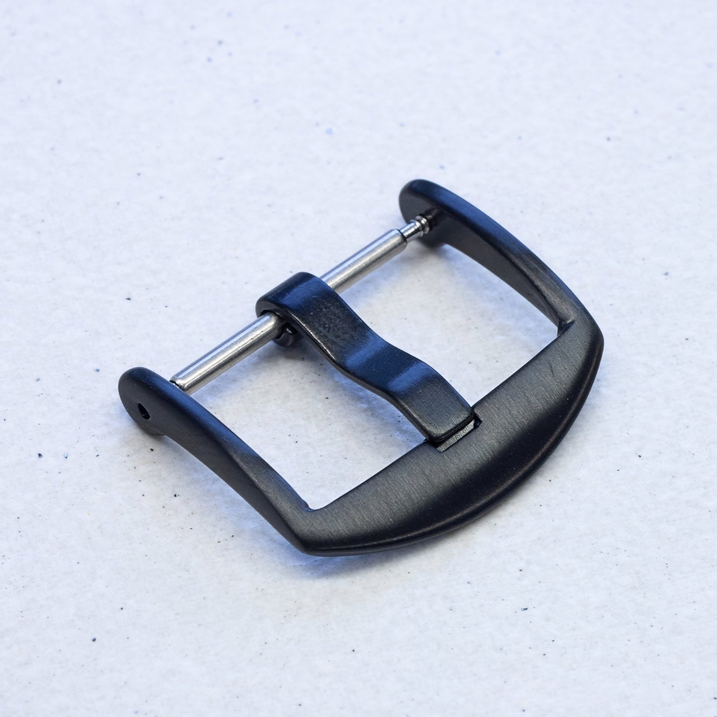 A black metal watch buckle with a brushed finish and silver-toned spring bars, shown on a light textured background.
