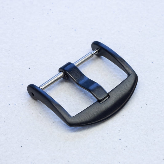 A black metal watch buckle with a brushed finish and silver-toned spring bars, shown on a light textured background.